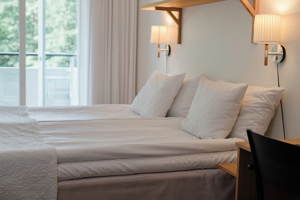 A hotel room with a large bed, two wall-mounted lamps, and a view of greenery outside.