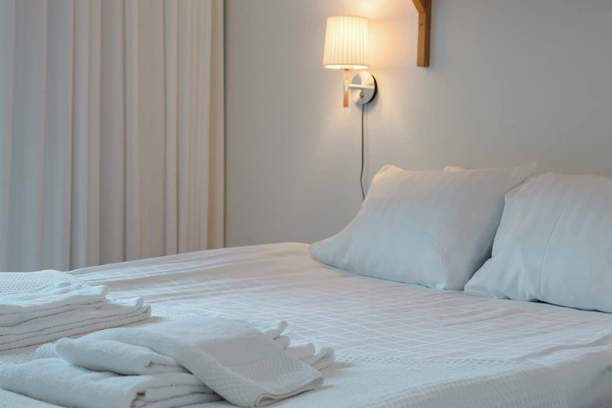 A neatly made bed with white linens and pillows, a wall-mounted lamp, and a window with blinds.