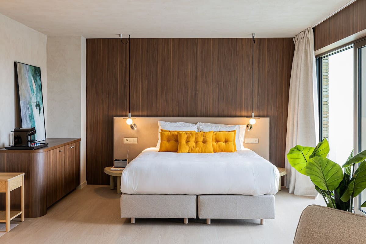 A modern hotel room with a large bed, a bedside table, a desk, and a large window with curtains.