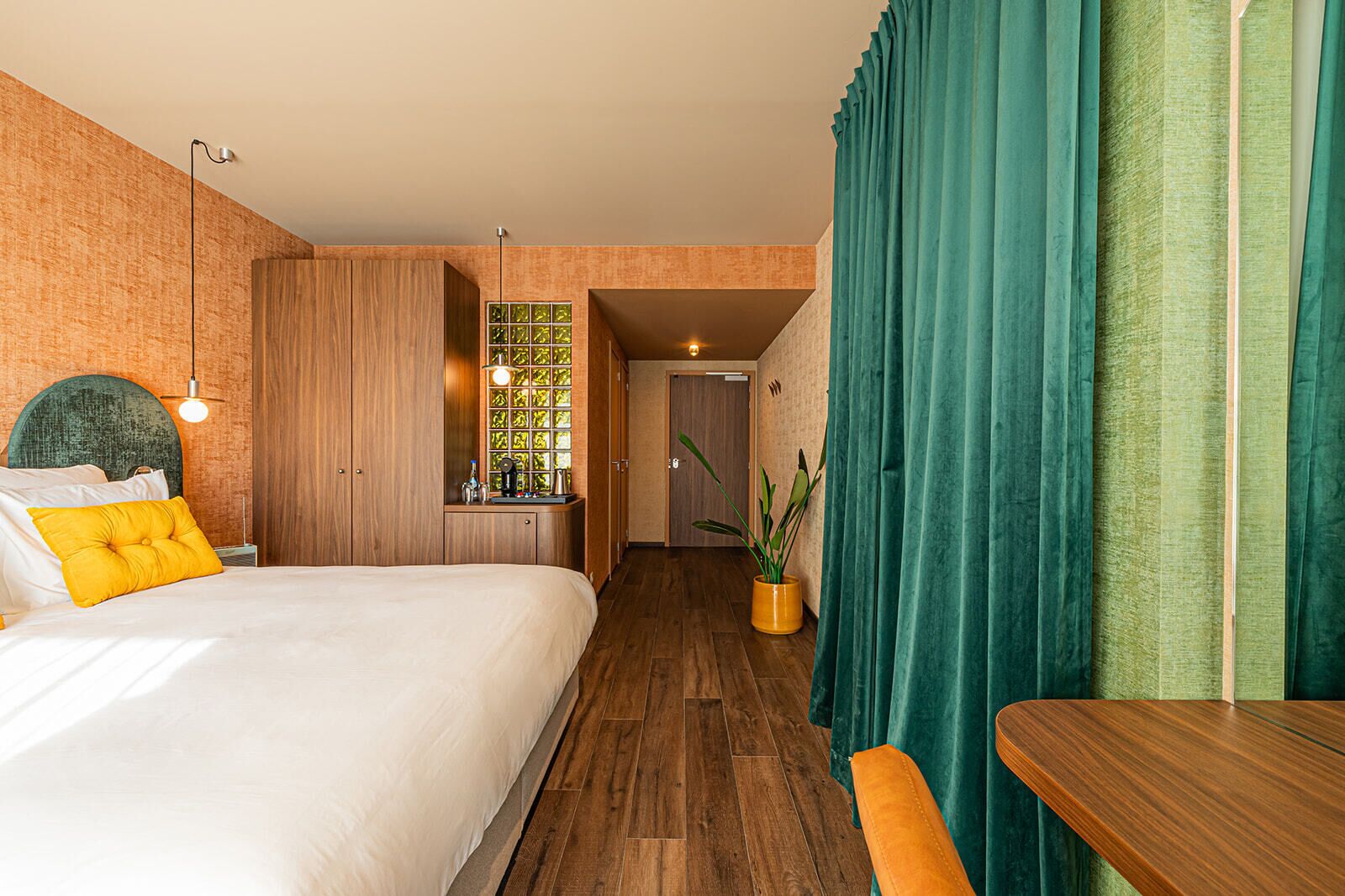 A hotel room with a bed, a desk, a potted plant, and a window with curtains.