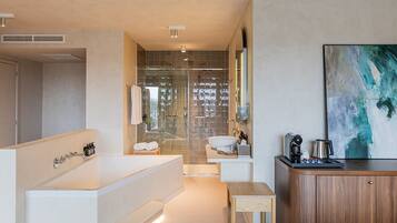 Een moderne badkamer met een ruim bad, een douche en een wastafel. Er is een houten kastje en een klein tafeltje met toiletartikelen.