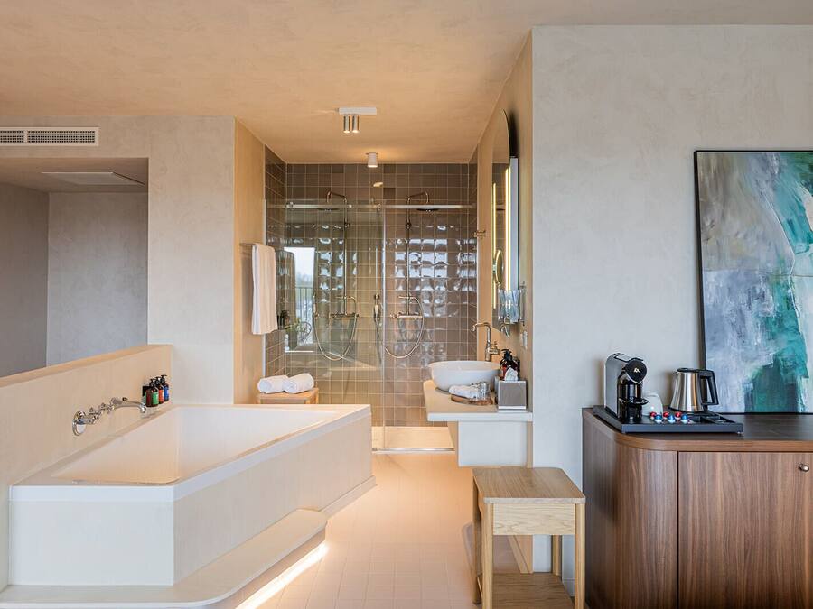Een moderne badkamer met een ruim bad, een douche en een wastafel. Er is een houten kastje en een klein tafeltje met toiletartikelen.