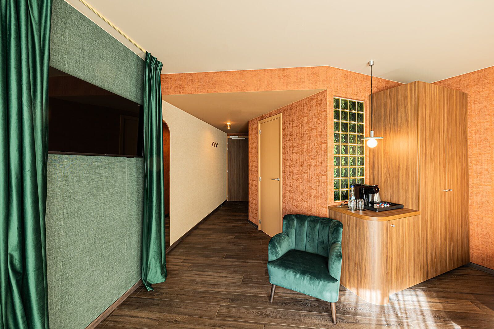 A hallway with a wooden floor, a green velvet armchair, a wooden counter with a coffee machine, and a door with a green glass window.