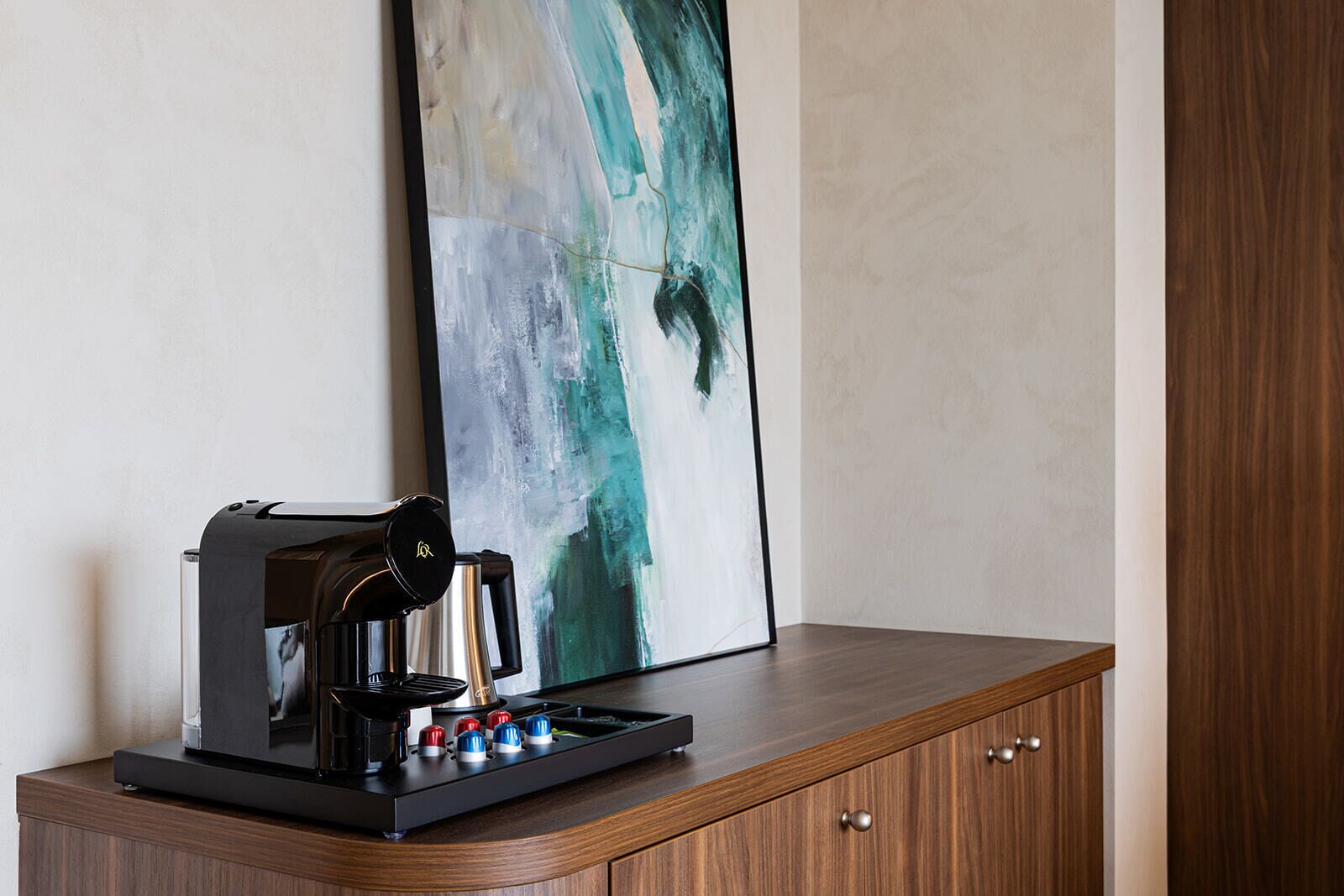 Una cafetera sobre un mueble de madera, con un cuadro abstracto minimalista de fondo.