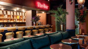 Eine Barbereich mit einer Neonreklame mit dem Schriftzug "The Bar", ein Bücherregal mit verschiedenen Gegenständen und Sitzgelegenheiten, darunter ein grünes Sofa und runde Tische mit dekorativer Beleuchtung.