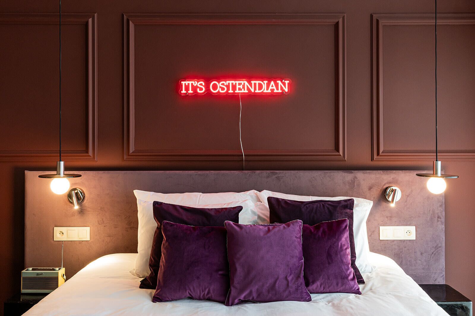 A hotel room with a bed, purple pillows, a neon sign reading "IT'S OSTENDIAN," and pendant lights.
