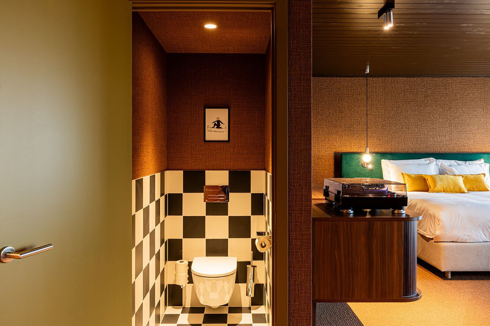 A hotel bathroom with a checkered tile wall, a toilet, and a bed with a green headboard and yellow pillows.