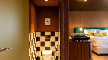A hotel bathroom with a checkered tile wall, a toilet, and a bed with a green headboard and yellow pillows.