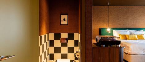 A hotel bathroom with a checkered tile wall, a toilet, and a bed with a green headboard and yellow pillows.
