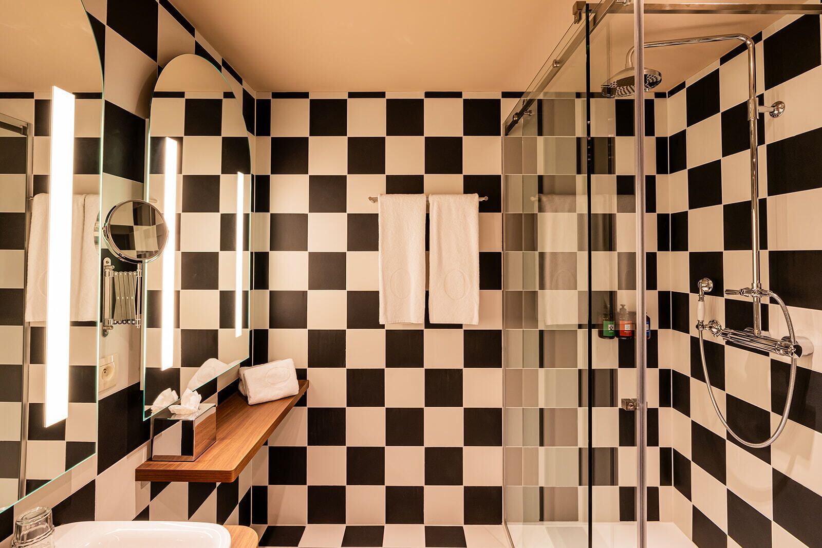 Un baño moderno con suelo de baldosas blancas y negras formando un diseño de cuadros, cabina de ducha de vidrio, estante de madera con artículos de aseo y espejo con marco redondo.