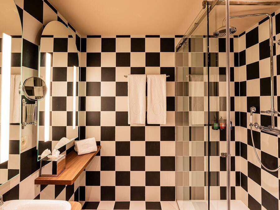 Une salle de bain moderne avec un carrelage noir et blanc à motifs géométriques, une douche en verre, une étagère en bois où sont rangés les produits de toilette et un miroir à cadre rond.