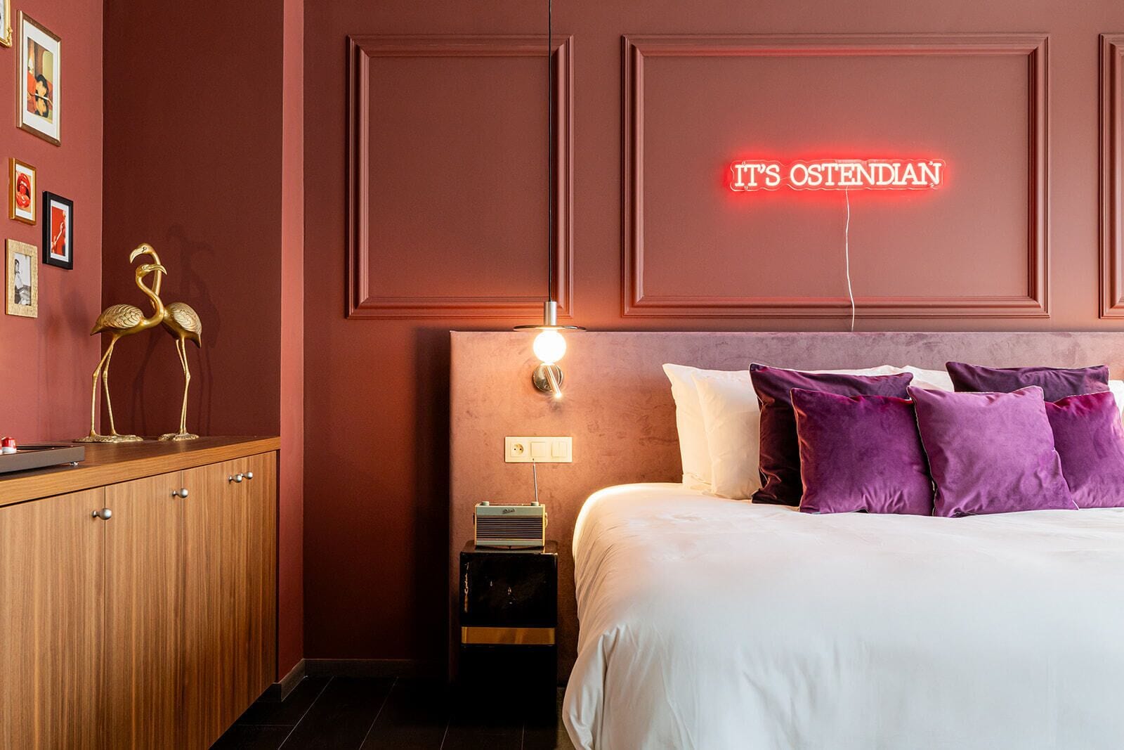Une chambre d’hôtel avec un lit, une table de chevet, une lampe, un panneau lumineux au néon et une commode en bois ornée d’œuvres d’art.