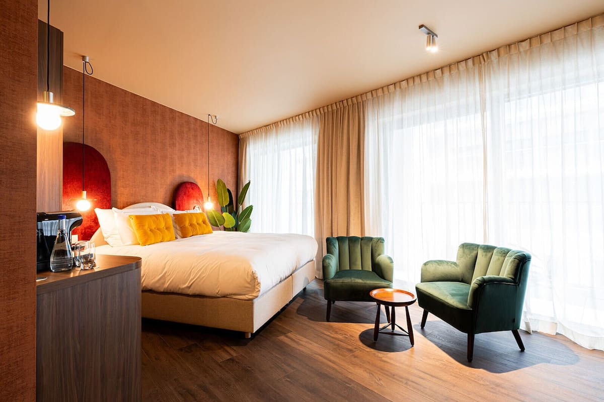 A hotel room with a bed, two armchairs, a small table, a bedside table with a lamp, and a large window with curtains.