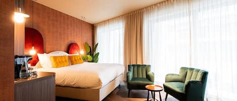 A hotel room with a bed, two armchairs, a small table, a bedside table with a lamp, and a large window with curtains.