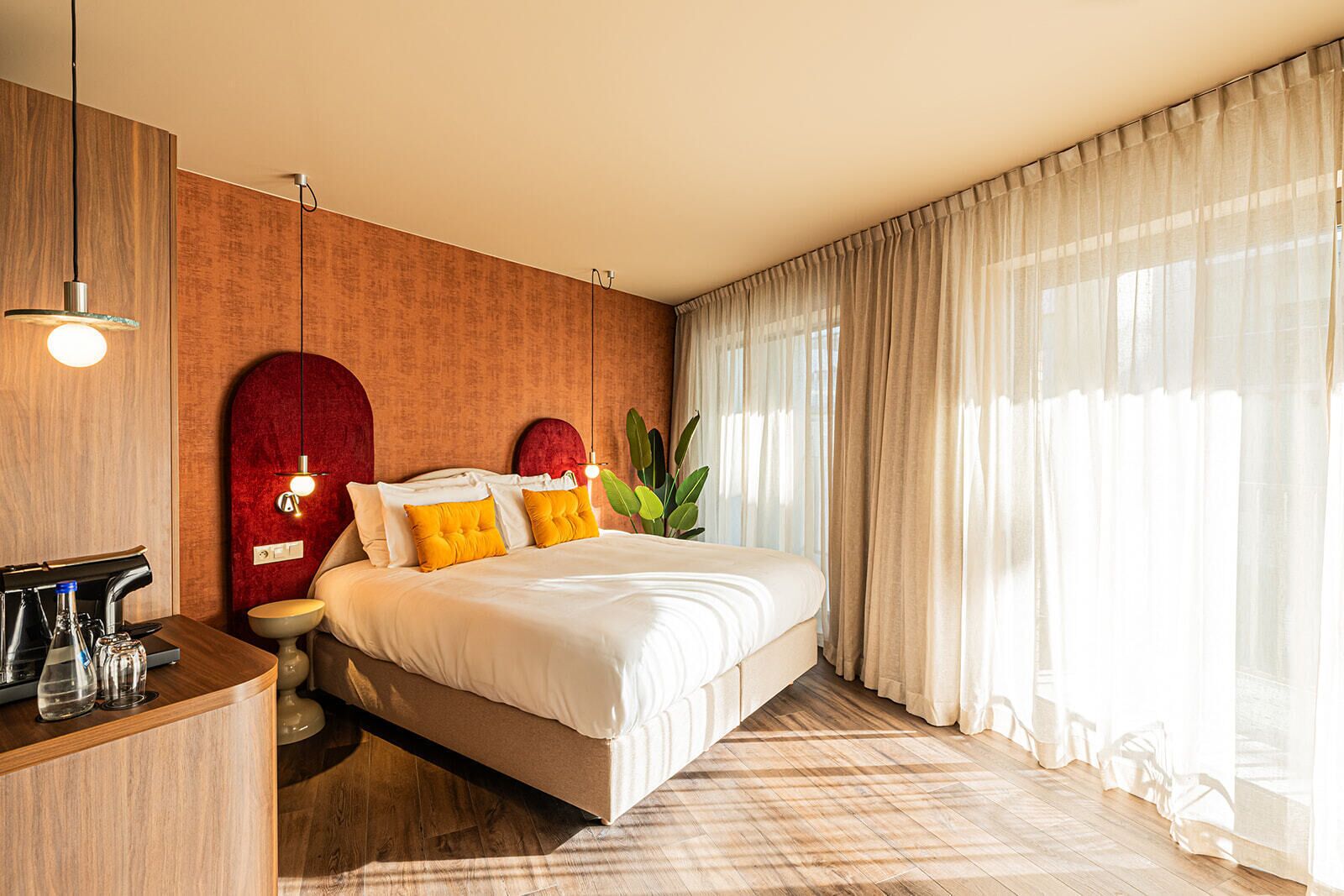 Ein modernes Hotelzimmer mit einem Holz-Kopfteil, einem Nachttisch mit Kaffeemaschine, einem großen Bett mit weißen und gelben Kissen sowie transparenten Vorhängen, die natürliches Licht hereinlassen.