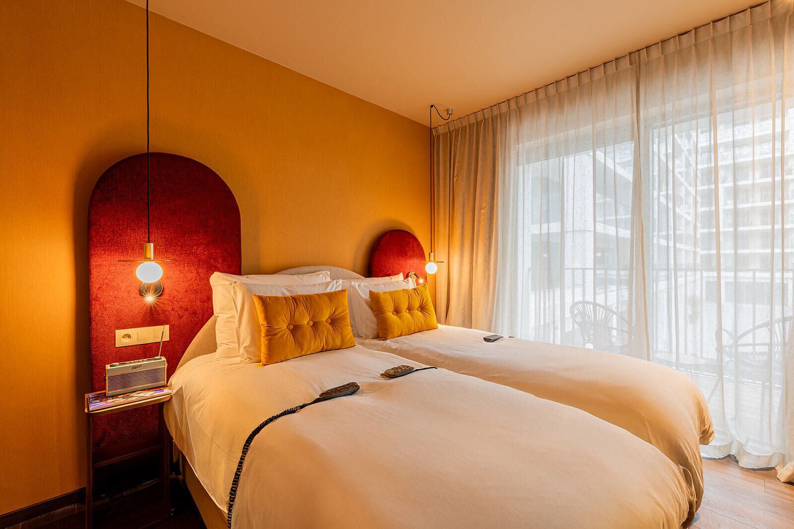 A hotel room with two beds, a red accent wall, a large window with sheer curtains, and a small table with a lamp and a remote control.