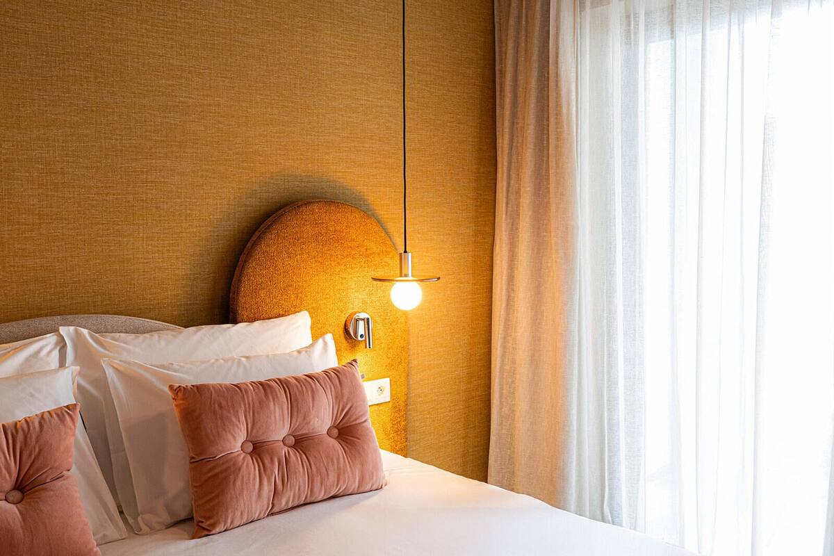 A hotel room with a bed, pillows, a wall-mounted light fixture, and a window with curtains.