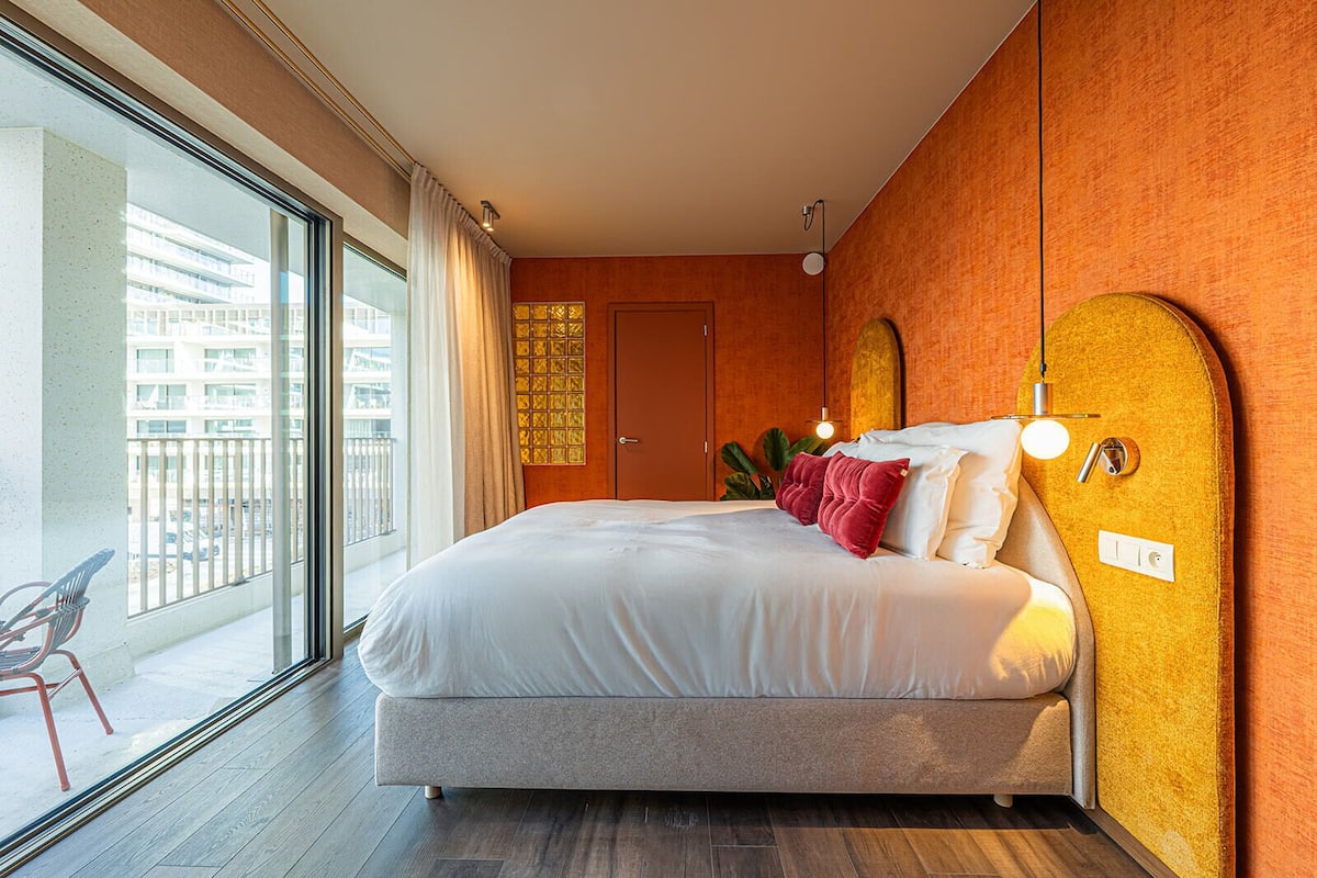A modern hotel room with a large bed, a balcony with a chair, and a view of the cityscape.
