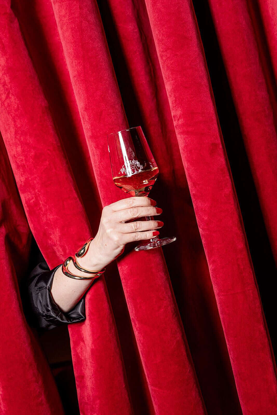 Una mano sosteniendo una copa de vino tras cortinas rojas.