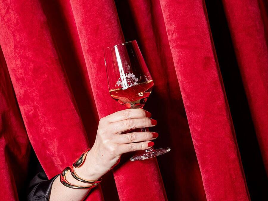 A hand holding a wine glass behind red curtains.
