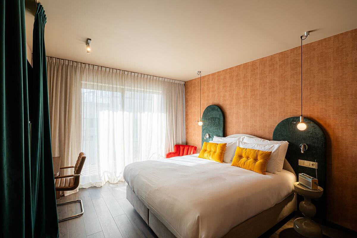 A hotel room with a large bed, a chair, a bedside table with a lamp, and a window with curtains.