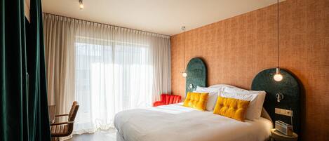Habitación de hotel con una cama grande, una silla, una mesita de noche con lámpara y un ventanal con cortinas.