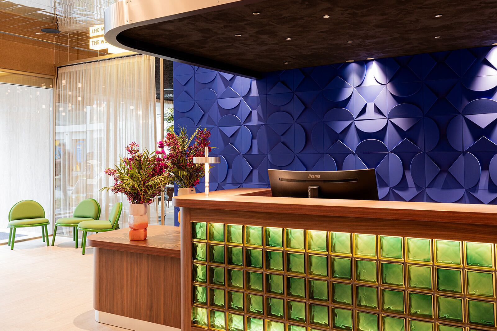 A reception desk with a blue geometric wall design, a wooden counter, and green chairs.