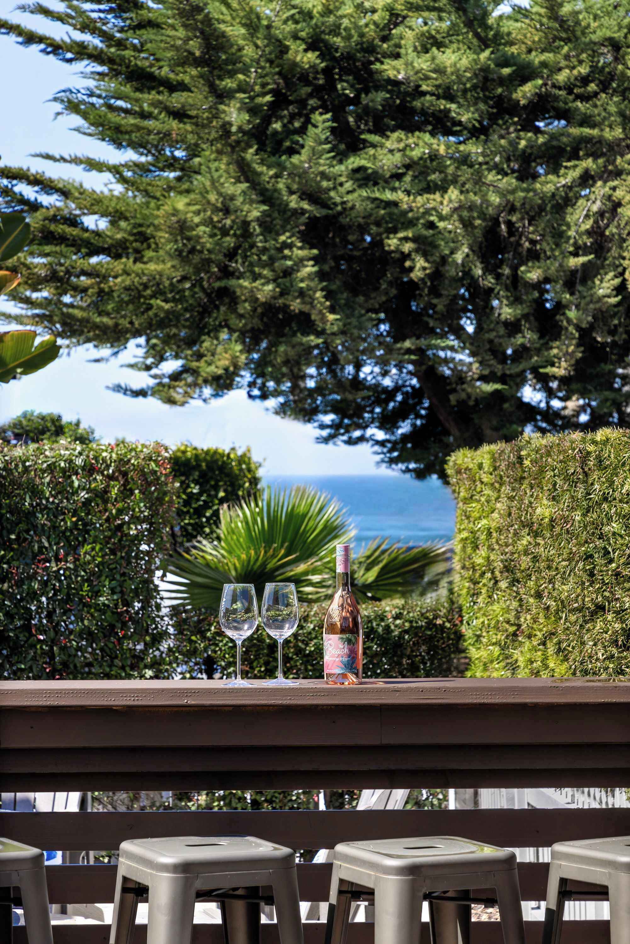 A table with two glasses and a bottle, set against a backdrop of lush greenery and a view of the ocean.