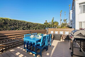 Un patio in legno con un tavolo blu apparecchiato per mangiare, un barbecue e vista su palme e un edificio sullo sfondo.