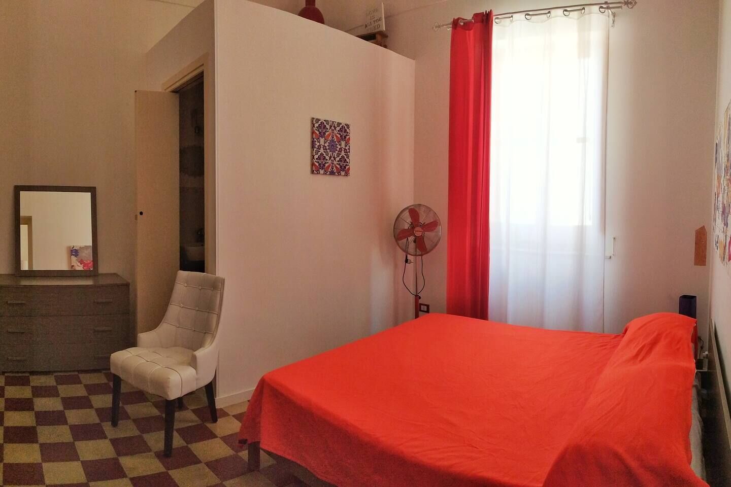 Een slaapkamer met een geruite vloer, een bed met een rode deken, een stoel, een kapstok, een ventilator en een raam met rode gordijnen.