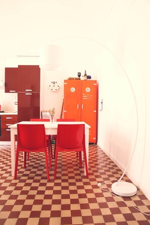 Une pièce avec un sol à carreaux, une table blanche avec des chaises rouges, un meuble orange et un lampadaire blanc.