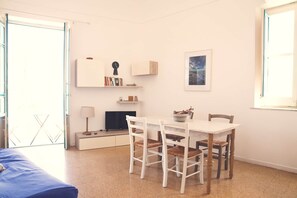 Una habitación con cama, una mesa de comedor con sillas, televisor, estantería y ventana.