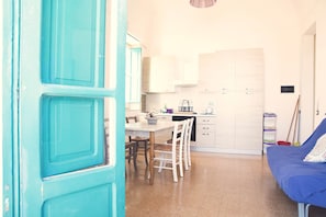 Cocina compacta con armarios blancos, una mesa de comedor con sillas y un sofá azul en una habitación luminosa.