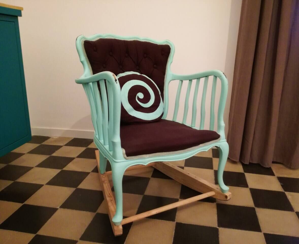 Een smaragdgroene schommelstoel met een bordeauxrode kussen en een spiraalvormig kussen, geplaatst op een geruite vloer.