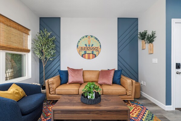 A living room with a tan sofa, blue armchair, and a coffee table with a plant. - Branson Breeze: 4BR Family Retreat with Arcade & 5★ Location, Double Living Room (Branson)