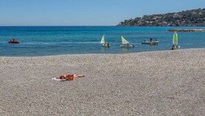 Una playa de guijarros con varios veleros en el agua, una costa lejana con edificios, y una persona tomando el sol.
