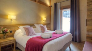 A hotel room with a large bed, wooden headboard, two bedside lamps, a nightstand with a vase of flowers, and a window with curtains.