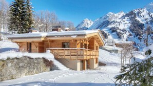 A wooden chalet with a balcony, surrounded by snow-covered mountains and trees.