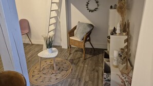 A room with a wooden floor, a wicker chair with a cushion, a small round table with a plant, a ladder leaning against the wall, and a shelf with decorative items. - Studio 2/4 pers near center and cable car (Cauterets)