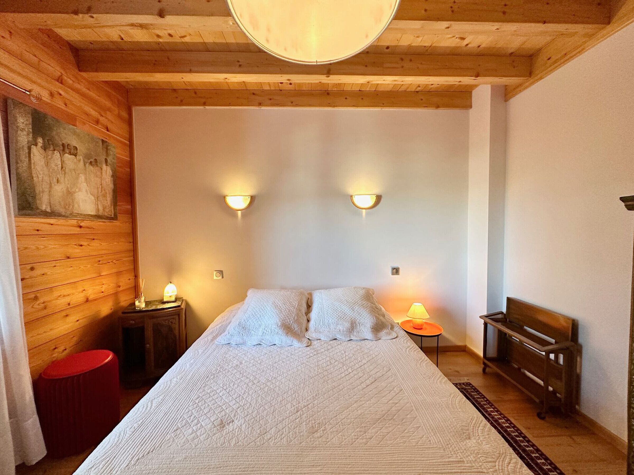 Ein Schlafzimmer mit einem großen Bett, zwei Wandlampen, einer hölzernen Bank und einem roten Hocker.