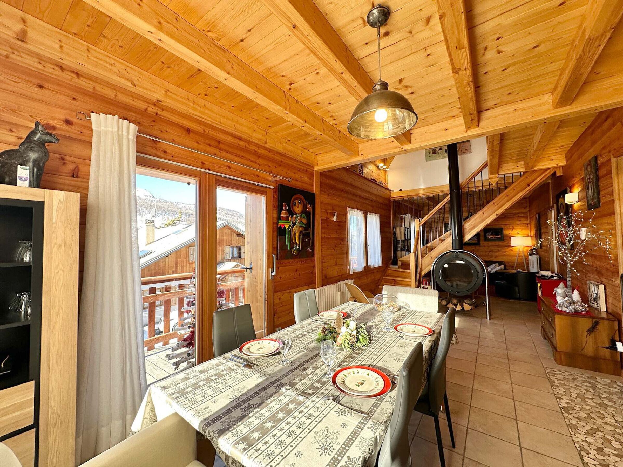 Interno di una baita in legno con un tavolo da pranzo apparecchiato per quattro persone, una scala e vista all'esterno.