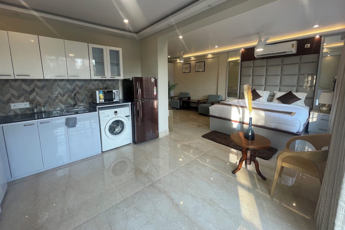 A modern hotel room with a bed, a small seating area, a kitchenette, and a washing machine.