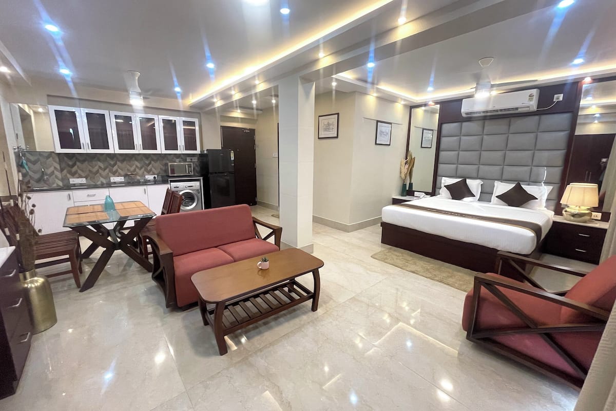 A modern hotel room with a bed, a sofa, a dining table, and a kitchen area.
