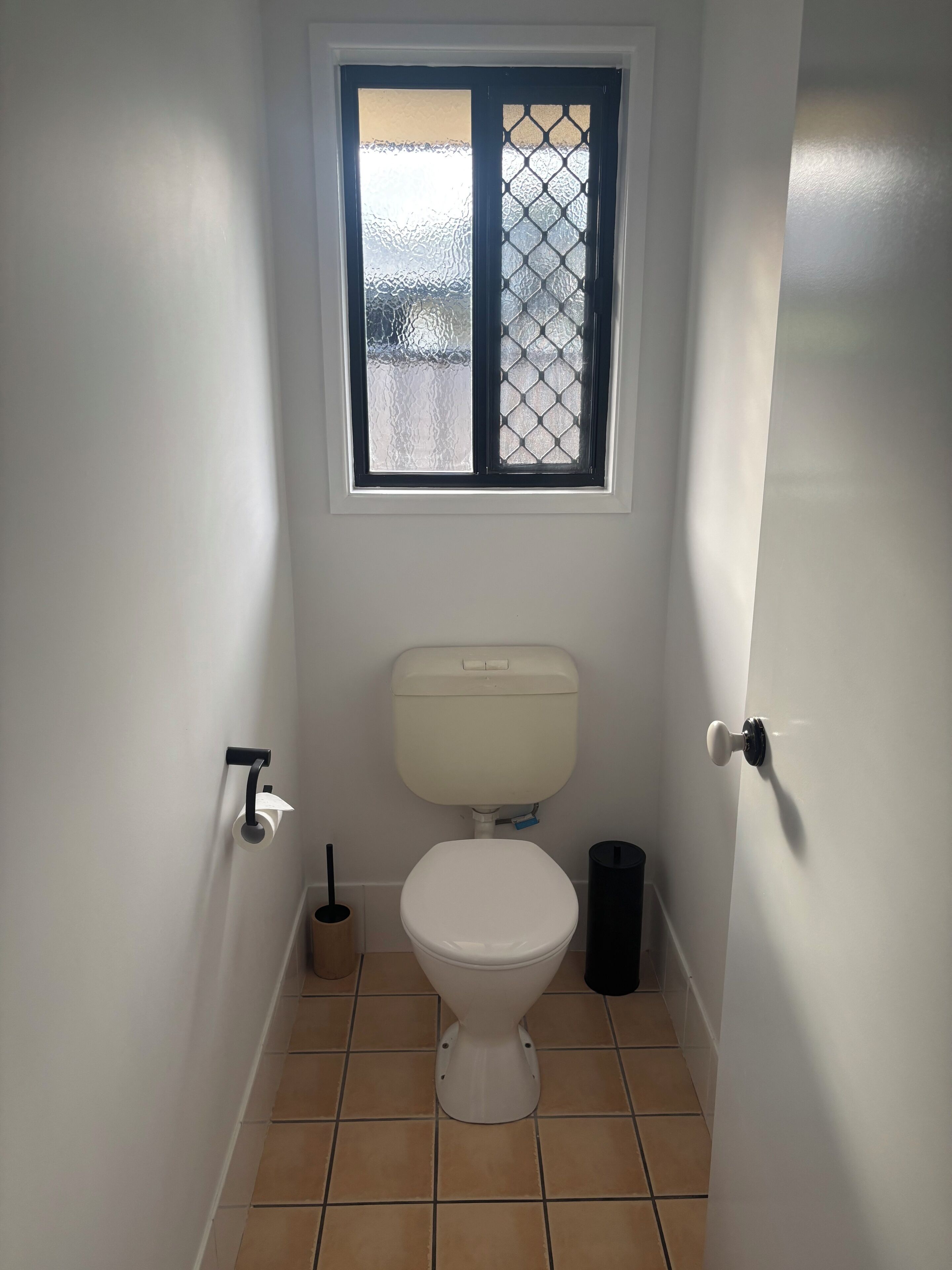 A small, narrow bathroom with a toilet, a window with a metal grid, and a trash bin.