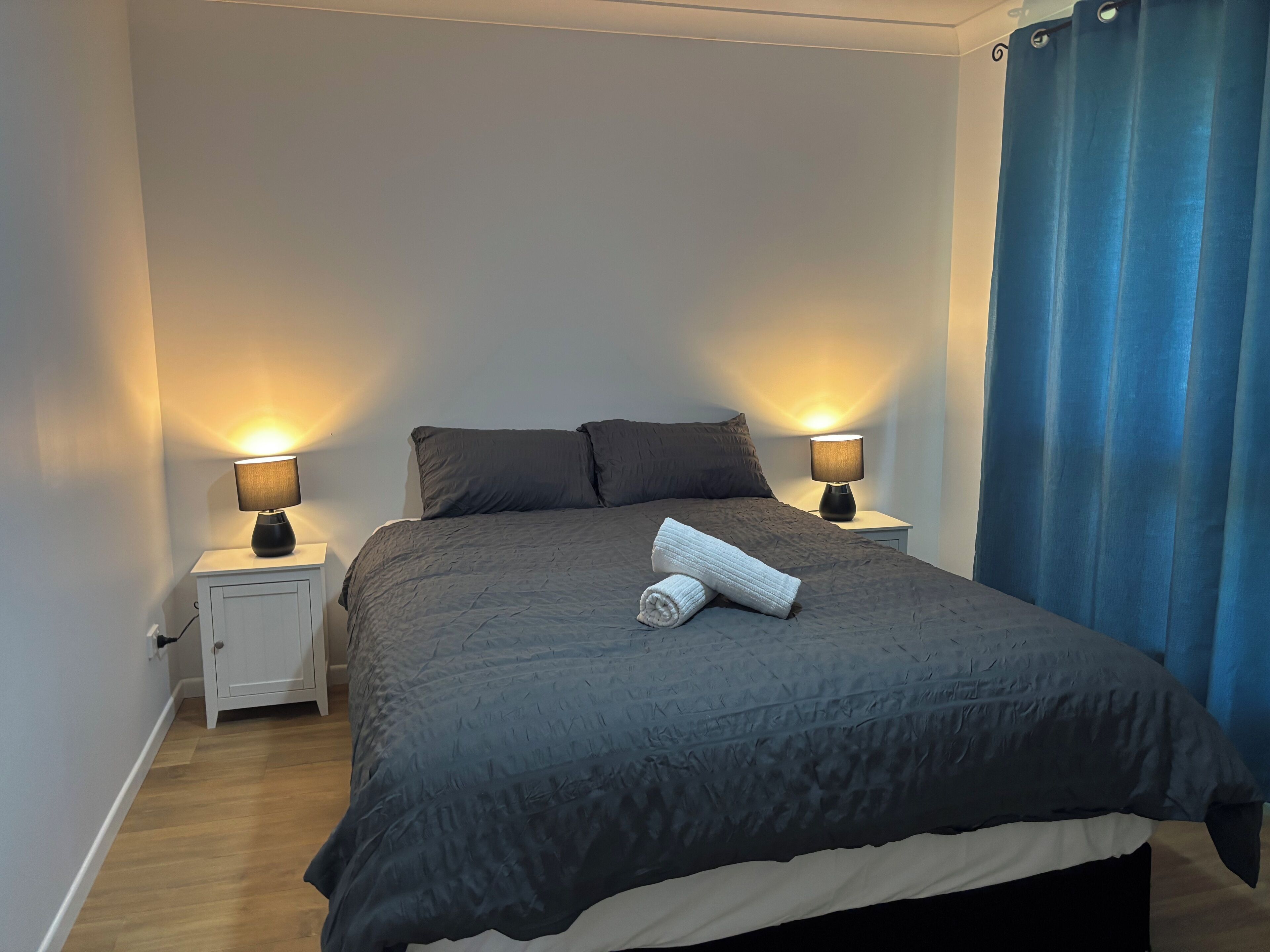 A neatly made bed with a dark bedspread, two bedside lamps, and a rolled towel on the bed.