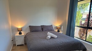 A neatly made bed with a grey quilt, two bedside lamps, and a window with a view of greenery.