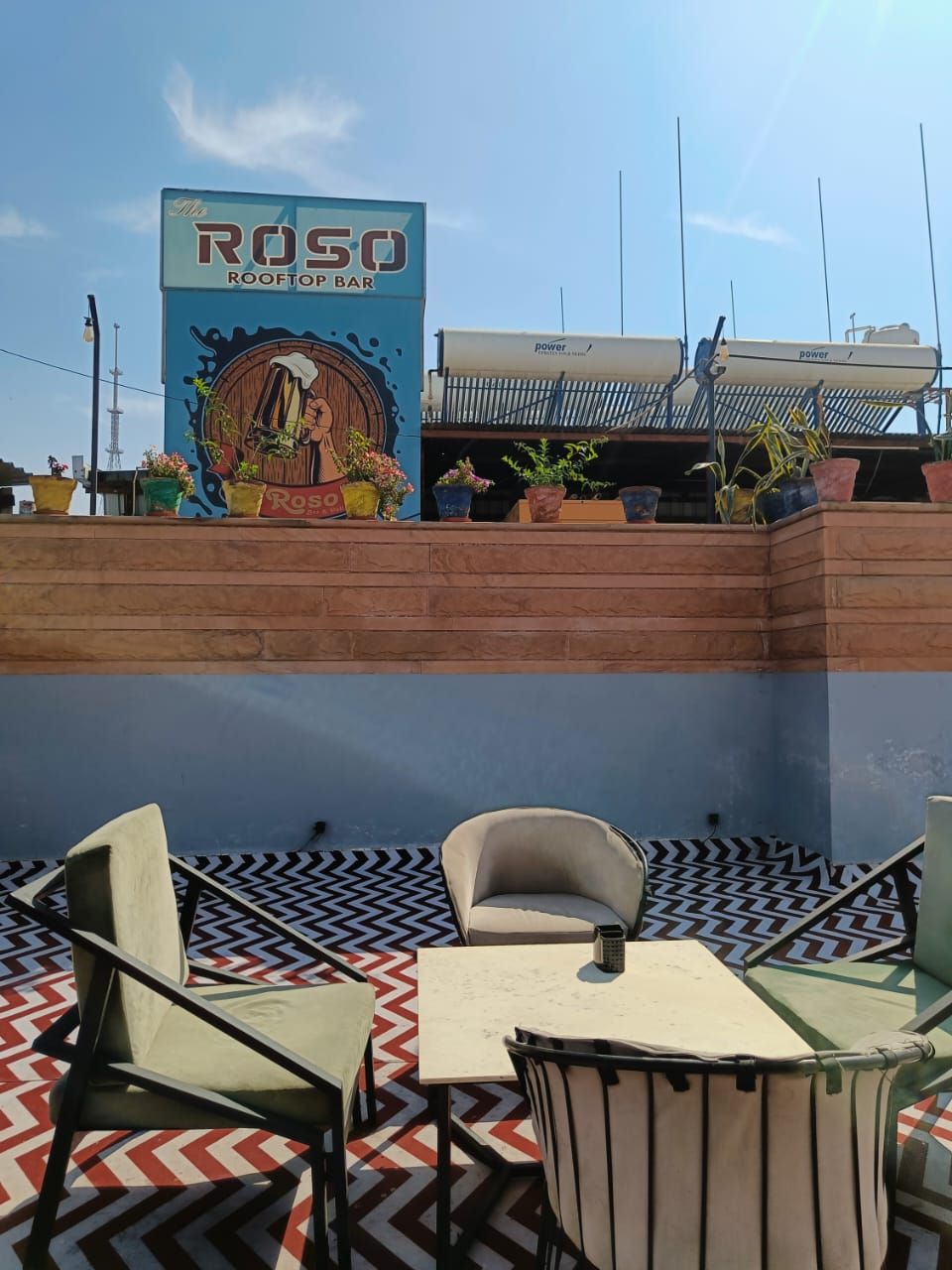 An outdoor seating area with a 'RósO ROOFTOP BAR' sign, potted plants, and a patterned floor.