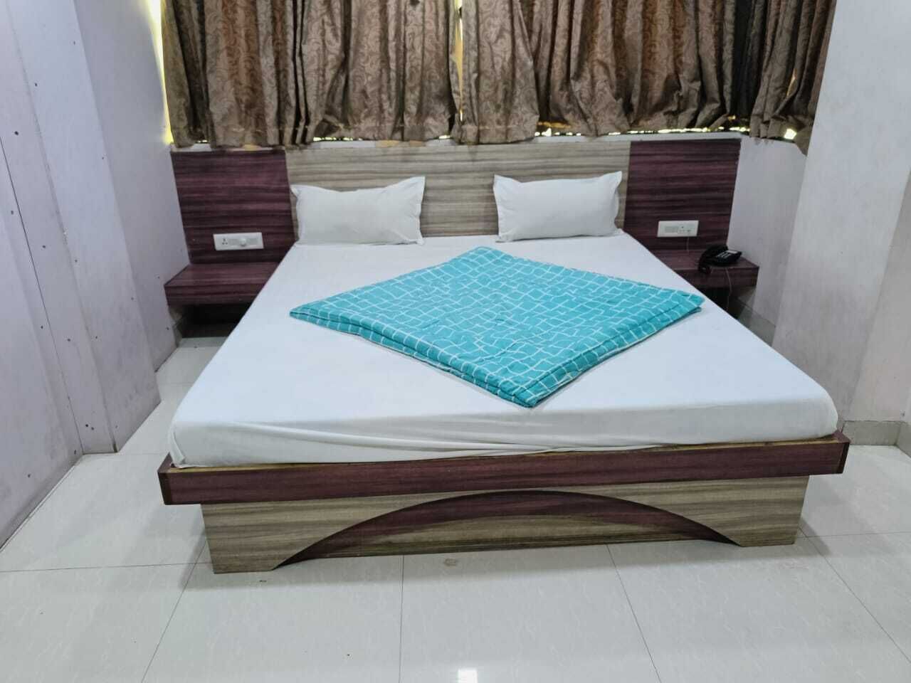 Una cama bien tendida con colchón blanco y colcha estampada en color verde azulado, flanqueada por dos mesitas de noche de madera con lámparas blancas.