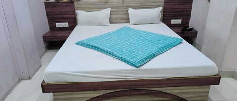 Una cama bien tendida con colchón blanco y colcha estampada en color verde azulado, flanqueada por dos mesitas de noche de madera con lámparas blancas.