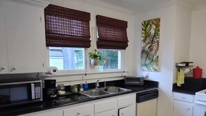Una cocina con armarios blancos, encimera negra, microondas, fregadero y estufa. Hay dos ventanas con persianas marrones, una planta en el alféizar, y un suelo a cuadros.
